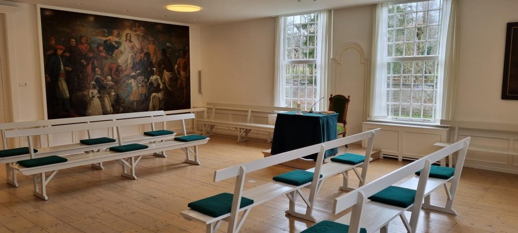 De Kleine Zaal met witte banken met groene kussens, hoge ramen en een groot historisch schilderij